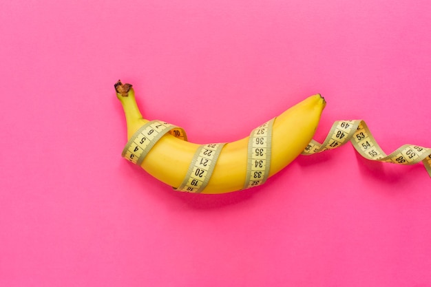 Banana gialla con nastro di misurazione su sfondo rosa. Concetto di dimensione del pene degli uomini. Lay piatto, vista dall'alto, copia spazio.