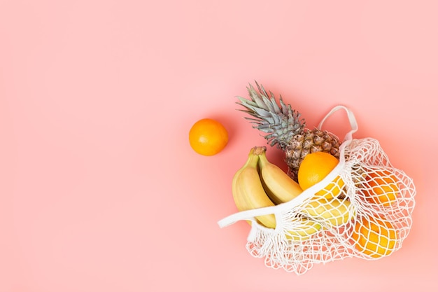 Banane, arance e ananas nella borsa della spesa, la borsa della stringa giacciono su sfondo rosa pastello Concetto di cibo