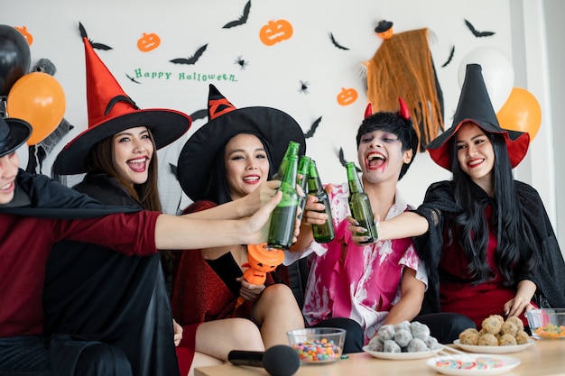 Banda di giovani asiatici in costume da strega, mago con festeggiare la festa di Halloween per una bottiglia tintinnante e un drink nella stanza. Gruppo teenager tailandese con festeggiare Halloween. Concetto di festa Halloween a casa.