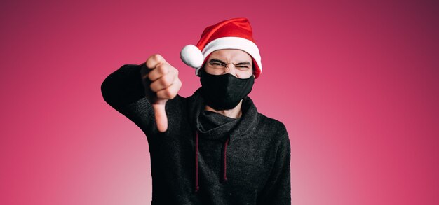 Banner di Natale giovane in maschera medica protettiva nera e cappello rosso mette antipatia con la mano