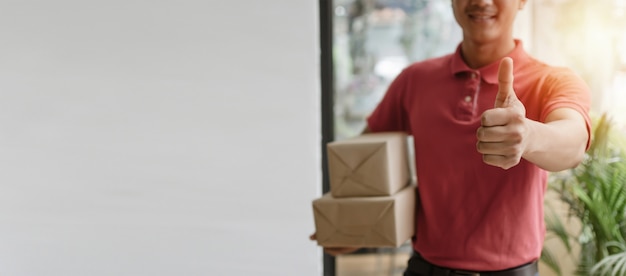Banner panoramico uomo di servizio di consegna in uniforme rossa che mostra i pollici in su e che tiene pronta casella di pacco postale inviare al cliente a casa