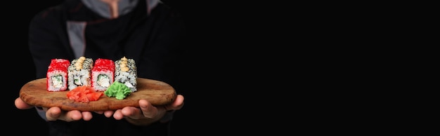 Banner primo piano delle mani di uno chef uomo in uniforme nera che tiene un vassoio in legno piatto pieno di sushi