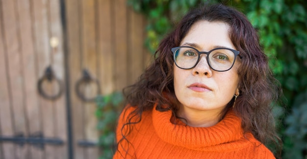 Banner Ritratto autunnale di una giovane donna attraente in occhiali eleganti in un maglione arancione alla moda lavorato a maglia su sfondo di foglie e grande spazio copia porta La ragazza cammina in città