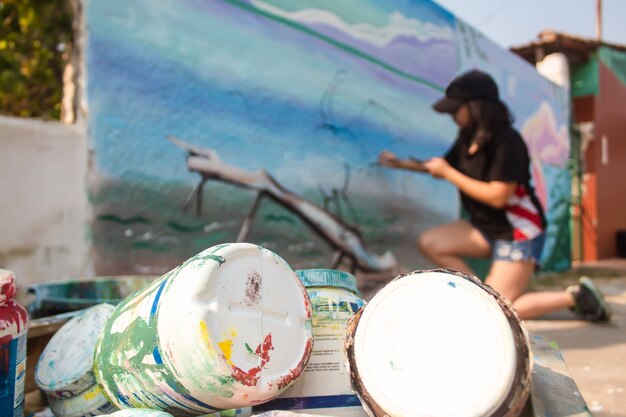 Barattoli di vernice impilati con una ragazza che dipinge un muro sullo sfondo.