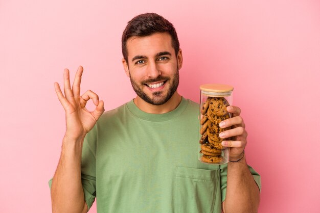 Barattolo di biscotti della holding del giovane uomo caucasico isolato su sfondo rosa allegro e fiducioso che mostra gesto giusto.