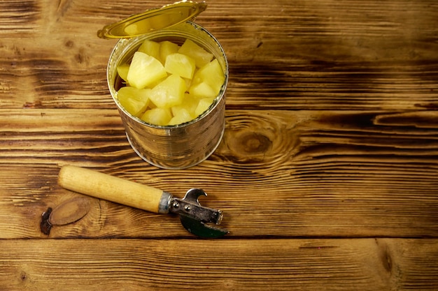 Barattolo di latta aperto di pezzi di ananas e apriscatole sul tavolo di legno