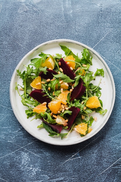 Barbabietole, arancia, pinoli, olio d'oliva, formaggio feta e insalata di rucola nel piatto di ceramica sul vecchio fondo della tavola di cemento