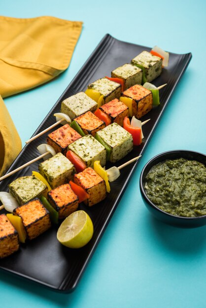 Barbecue Paneer Tikka Kabab - cubetti di ricotta indiana Tandoori in salsa rossa e bianca in bastoncini, serviti in un piatto con chutney verde su un tavolo colorato o in legno, messa a fuoco selettiva