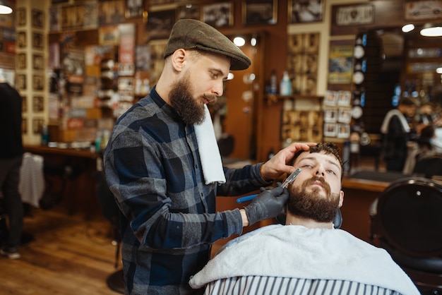 Barbiere con il rasoio, taglio della barba della vecchia scuola. Il barbiere professionale è un'occupazione alla moda. Parrucchiere maschio e cliente nel salone di parrucchiere