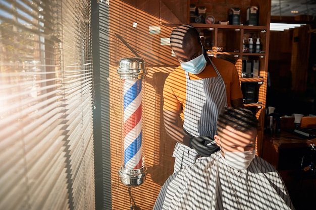 Barbiere in maschera medica che fa taglio di capelli per uomo nel negozio di barbiere