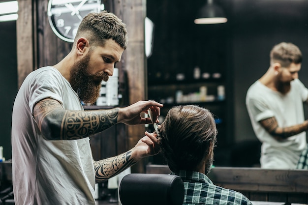 Barbiere professionista con braccia tatuate che tiene un pettine e che fa il taglio di capelli per il suo cliente