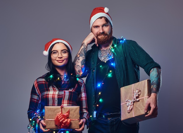 Barbuto tatuato hipster maschio e sexy bruna femmina vestita con una camicia in pile tiene i regali di Natale su sfondo grigio.