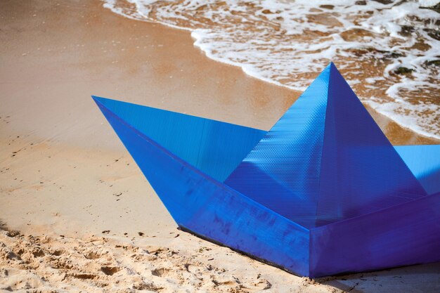 Barca di carta blu origami sulla spiaggia sabbiosa per il concept design, bellissima nave di carta a terra. Oggetto d'arte moderna, primo piano, sfondo oceano. Divertimento estivo all'aperto, viaggi, concetto di vacanza estiva