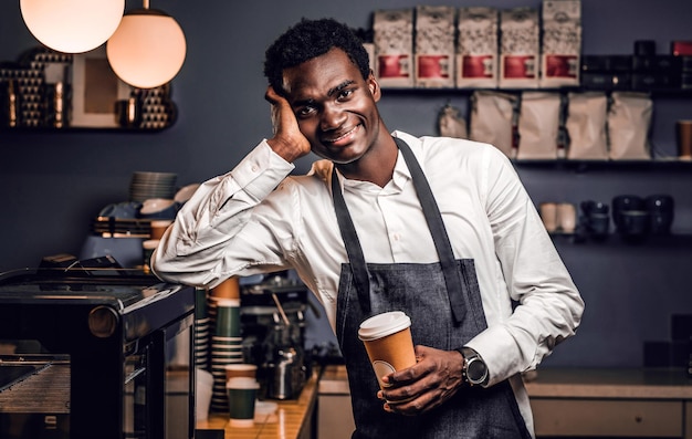 Barista africano stanco che tiene una tazza di caffè mentre si appoggia su un bancone in una caffetteria e guarda una macchina fotografica con uno sguardo felice.