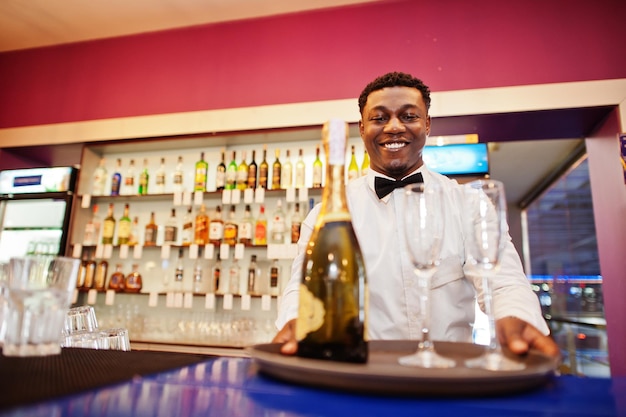 Barista afroamericano al bar che tiene champagne con bicchieri sul vassoio