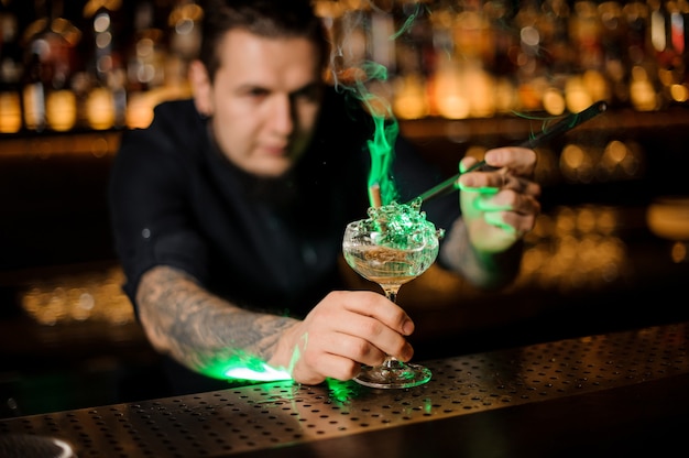 Barista aggiungendo al bicchiere da cocktail con una cannella affumicata aromatica di arancia essiccata con una pinzetta sul bancone del bar