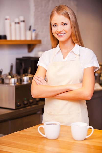 Barista allegro. Bella giovane barista femminile che tiene le braccia incrociate e sorride mentre si trova vicino al bancone del bar con due tazze di caffè vicino a lei