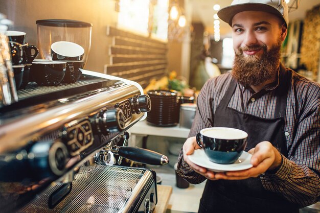 Barista allegro e piacevole che tiene tazza di caffè in mano. Sta in cucina alla macchina del caffè.