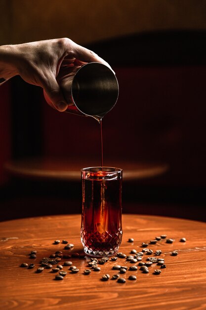 Barista che versa un cocktail da uno shaker in un bicchiere highball collins con una lancia di ghiaccio. I chicchi di caffè intorno su un tavolo di legno.