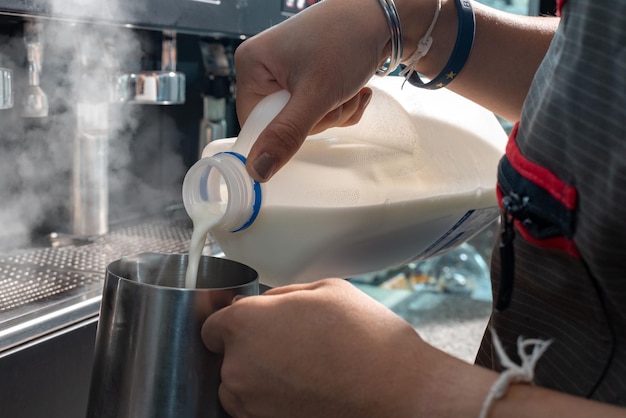 Barista donna del primo piano in grembiule che prepara e versa il latte nella tazza calda nella caffetteria