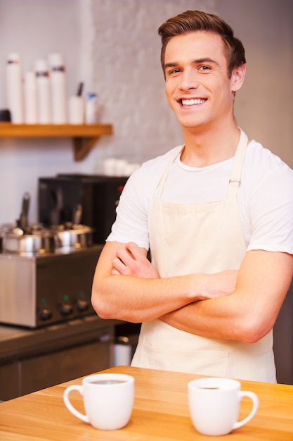 Barista fiducioso. Bel giovane barista maschio che tiene le braccia incrociate e sorride mentre si trova vicino al bancone del bar con due tazze di caffè vicino a lui