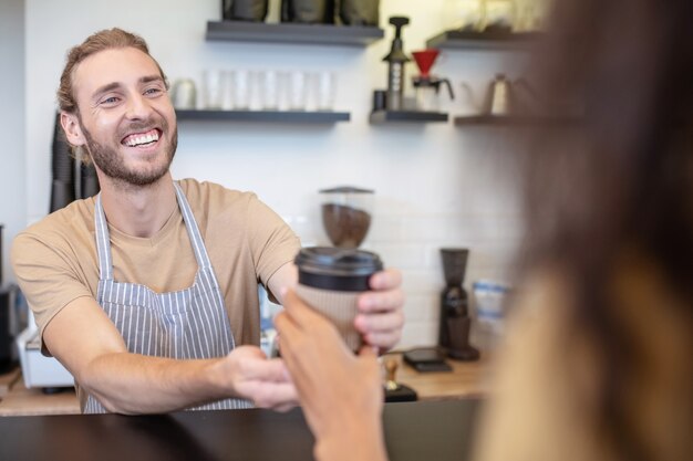 Barista gentile. Entusiasta giovane uomo barbuto con un sorriso a trentadue denti che dà caffè da asporto al cliente al bar