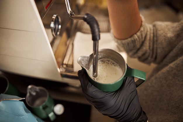 Barista in guanti di gomma che tiene una brocca con il latte e usa una lancia a vapore per mescolarlo