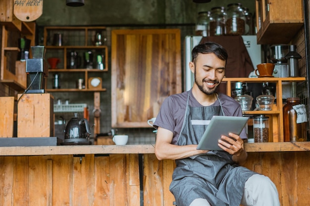 Barista maschio in grembiule che utilizza la tavoletta digitale mentre è seduto