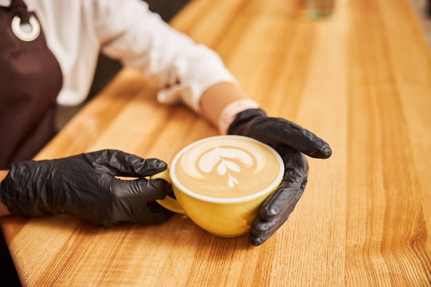 Barista non riconosciuto in guanti di gomma nera che mette una tazza di cappuccino su un bancone bar in legno