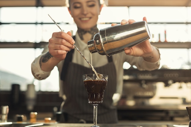 Barista tatuato felice che prepara cocktail Giovane ragazza che produce caffè freddo bevanda analcolica nel suo posto di lavoro. Concetto di piccola impresa, spazio di copia