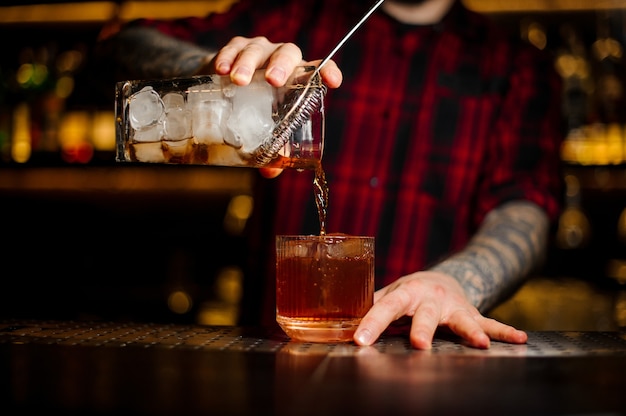 Barista versando un cocktail Old Fashioned dal misurino con colino a un bicchiere sul bancone del bar