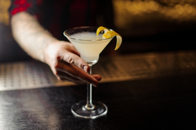 Barman che tiene un bicchiere di cocktail fresco acido e dolce decorato con buccia d'arancia sul bancone del bar