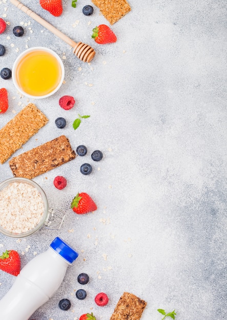 Barretta muesli di cereali biologici con frutti di bosco con cucchiaio di miele e barattolo di avena e bottiglia di bevanda al latte. Vista dall'alto. Fragola, lampone e mirtillo con mandorle noci.