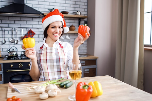 Basamento attraente adulto della donna in cucina e nella posa. Tenendo i peperoni in mano. Indossa un cappello festivo rosso. Festeggiamo Natale o Capodanno. Cucinare da solo.