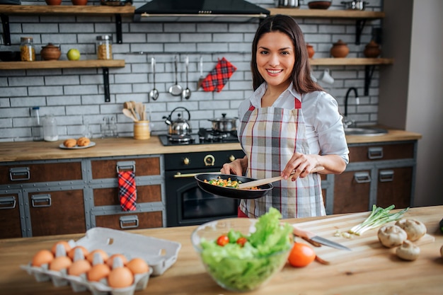 Basamento piacevole allegro della donna alla tabella in cucina e posa sulla macchina fotografica