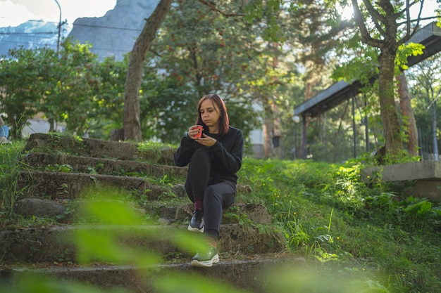 Basso angolo di giovane viaggiatore femminile in abbigliamento sportivo seduto su vecchie scale di pietra e bere caffè mentre riposa durante la passeggiata nel verde parco botanico