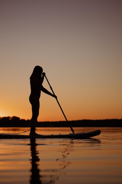 Basso angolo di silhouette donna in piedi su paddle board con pagaia in mano sul lago al tramonto con alberi sullo sfondo in estate Stile di vita attivo