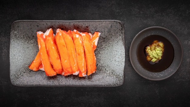 Bastoncini di granchio d'imitazione dell'Alaska con salsa di soia Wasabi e Shoyu in piatto di ceramica su fondo di struttura di tono scuro, vista dall'alto