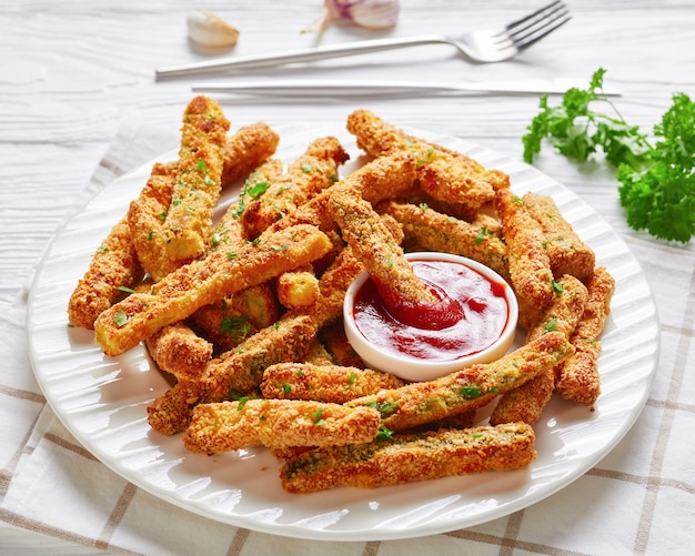 Bastoncini di zucchine impanate con pangrattato panko, parmigiano, spezie su una piastra bianca con ketchup su una tavola di legno, vista da sopra