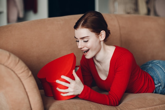 Beautifil giovane donna con i capelli rossi sdraiata nel suo divano in soggiorno e tenendo e aprire la confezione regalo a forma di cuore e sorriso. San Valentino o compleanno