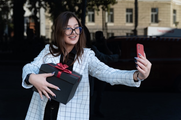 Beautiful stylish brunette with a gift in her hands makes a selfie on a smartphone.
