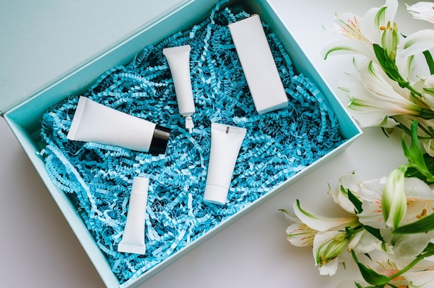 Beauty box con campioni di cosmetici per la cura del viso con trucioli di carta da imballaggio blu e alstroemeria bianca, vista dall'alto