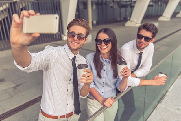 Bei giovani uomini d'affari e signora con gli occhiali da sole stanno tenendo il caffè facendo selfie utilizzando lo smartphone e sorridendo in piedi sul balcone dell'edificio per uffici