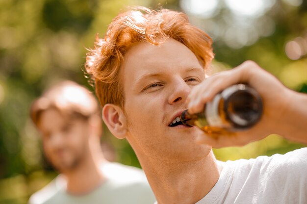 Bei momenti. Bel ragazzo dai capelli rossi che beve dalla bottiglia godendosi il momento e l'amico a distanza sulla natura in una giornata di sole estivo