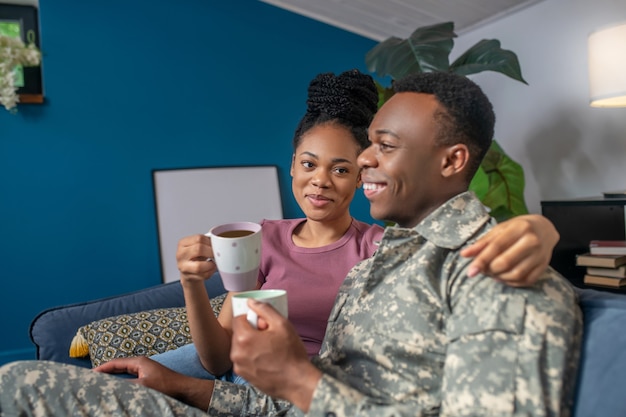 Bei momenti. Giovane militare dalla pelle scura in mimetica e bella moglie con l'acconciatura che beve caffè sul divano a casa di ottimo umore