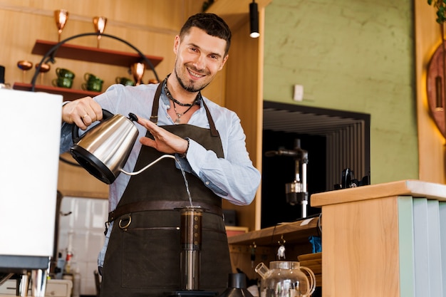 Bel barista versa acqua calda in aeropress con caffè e guarda a porte chiuse e sorride nella caffetteria