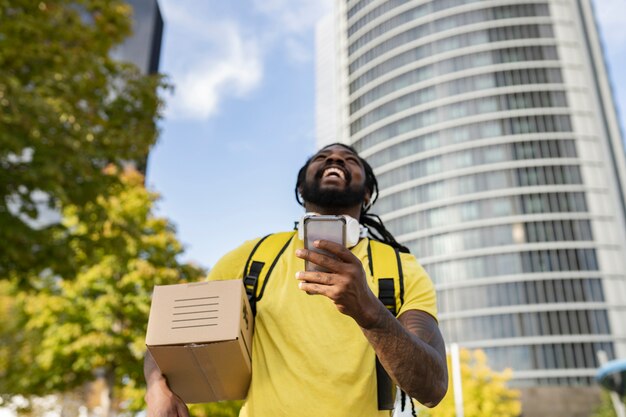 Bel brasiliano con i dreadlocks fattorino con una scatola, con le cuffie sorridenti guardando il suo smartphone per individuare, concentrarsi sulla faccia del telefono fuori fuoco