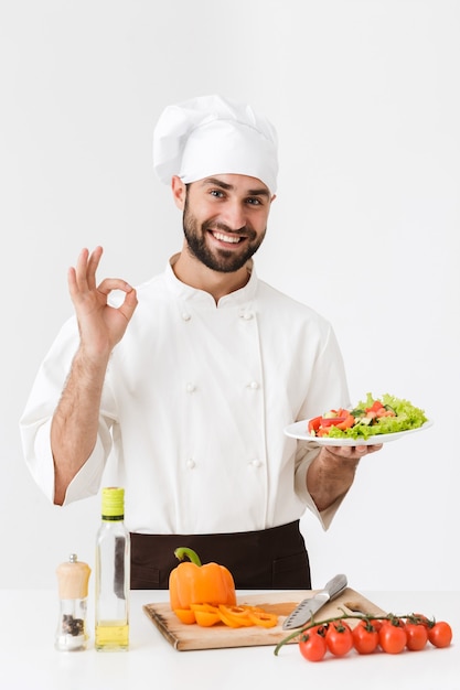 bel cuoco in uniforme che mostra un delizioso segno e tiene piatto con insalata di verdure isolato su muro bianco