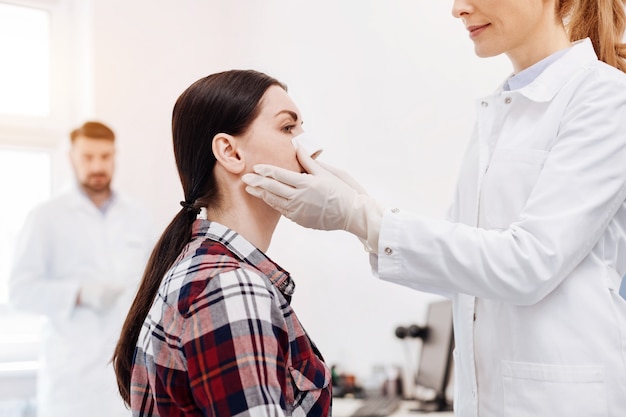 Bel dottore positivo allegro che tiene il viso dei suoi pazienti e mette una medicazione medica sul naso mentre si trova di fronte a lei