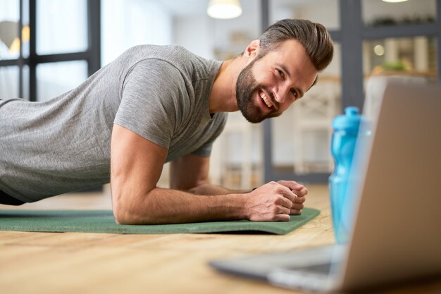 Bel gentiluomo barbuto che guarda l'allenamento online sul laptop e sorride mentre fa esercizio